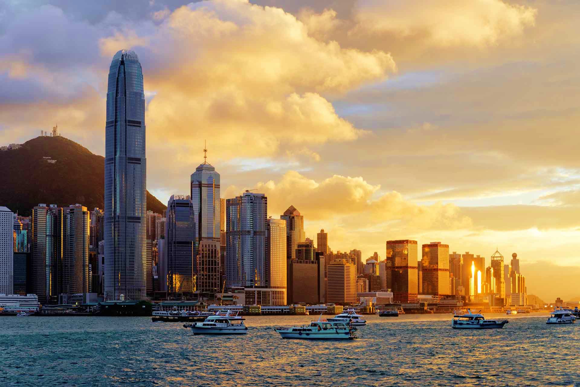 Hong Kong skyline at sunset with International Finance Centre tower, Victoria Harbour waterfront, and boats, golden sky with